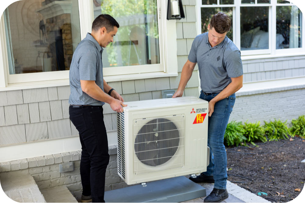 Two technicians install a Mitsubishi outdoor air conditioner unit on a mounting pad beside a house.